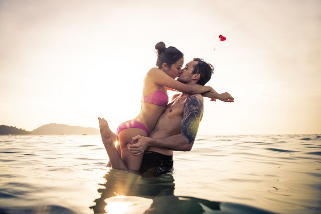 Couple of lovers kissing into water at sunset - Romantic scene of multiethnic couple