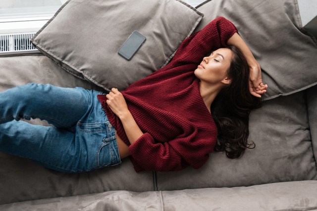 Top view of a smiling asian woman in sweater sleeping on a couch at home with blank screen mobile phone
