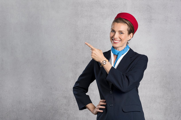 Portrait of air hostess pointing against grey background with copy space. Happy flight assistant indicating. Young flight attendant on gray background showing something while looking at camera.