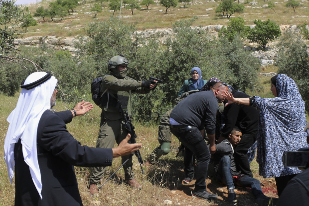 In this Thursday, Apri 18, 2019 photo, an Israeli soldier points his pistol at a group of Palestinians gathering around a wounded Osama Hajahjeh, 16, near the village of Tekoa, West Bank. Hajahjeh said was shot in his legs last week by Israeli soldiers while he was handcuffed and blindfolded, shortly after being arrested. The military said it was investigating the incident, which it said occurred as Palestinian youths were throwing stones at Israeli soldiers. (AP Photo/Mustafa Allbadan)
