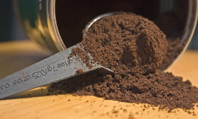 Coffee can lying on bright wood surface with scoop filled with coffee grounds with some spilling over.