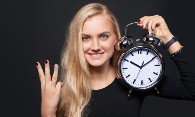 Hand counting - number three. Smiling woman holding big alarm clock showing three fingers, portrait over grey background