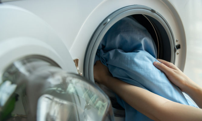 Woman’s hand loading dirty blue bed sheets in a white washing machine.