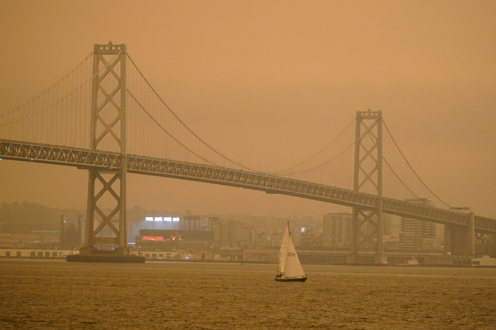 Under darkened skies from wildfire smoke, a sailboat makes its way past the San Francisco-Oakland Bay Bridge and lights at Oracle Park Wednesday, Sept. 9, 2020, in San Francisco. People from San Francisco to Seattle woke Wednesday to hazy clouds of smoke lingering in the air, darkening the sky to an eerie orange glow that kept street lights illuminated into midday, all thanks to dozens of wildfires throughout the West. (AP Photo/Eric Risberg)