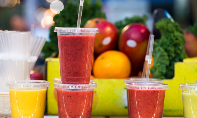 Close up color image depicting freshly made fruit juices and smoothies on display in a row and for sale at Spitalfields Market, a food and vintage market in east London, UK. In the background, defocused, is a pile of fresh fruit and vegetables - the raw ingredients used to make the smoothies. Room for copy space.