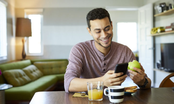 Smiling young man using smart phone at dining table. Male is having granny smith apple at home. He is enjoying social media and breakfast.