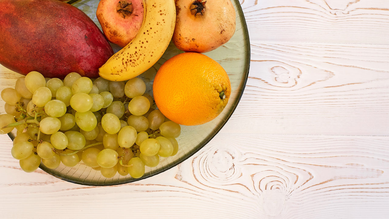 Juicy fruit grapes, banana, mango, pomegranate, oranges on a plate on a wooden white table. Copy space, flat lay.
