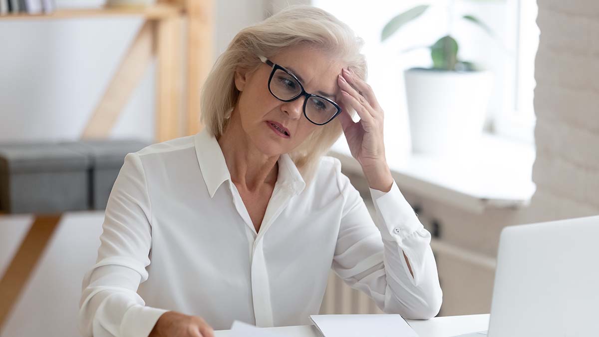 Pensive aged businesswoman in glasses sit at office desk thinking of problem solution, thoughtful senior woman worker in glasses look at laptop screen hold document pondering or making decision